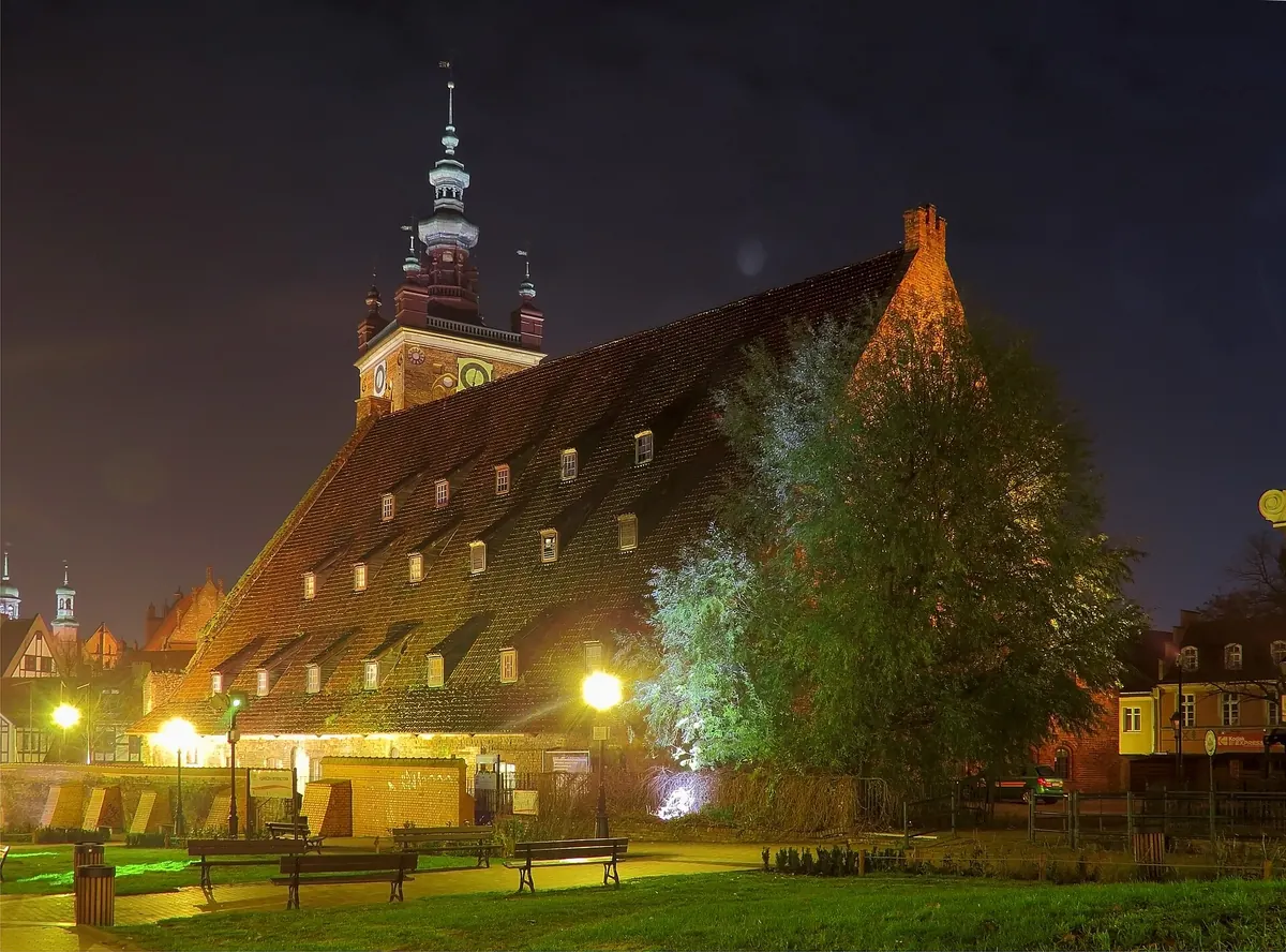 Obrovská strmá střecha Velkého mlýna v Gdaňsku nasvícená zespodu v noci, za ní se tyčí věž kostela sv. Kateřiny