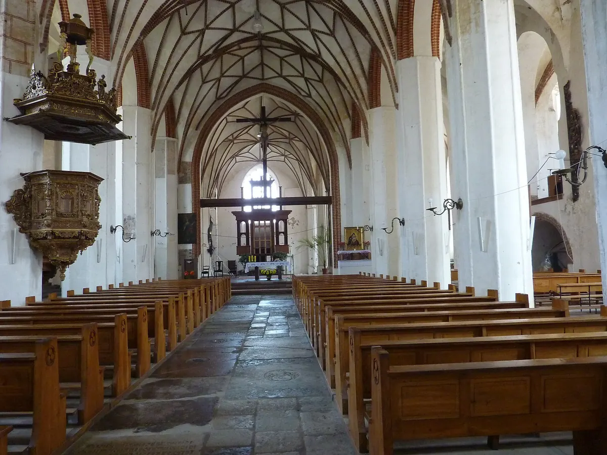 Dlouhá loď kostela sv. Kateřiny v Gdaňsku pohledem z boční lodi: řady dřevěných lavic, bílé pilíře s cihlovými žebry a pozlacená kazatelna vlevo